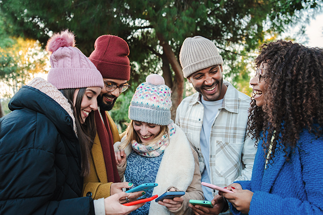 Group of multiracial young friends with coats and hats, smiling and watching the social media with a cellphone app. Portrait of happy people having fun sharing media and laughing with a smart phone. Communication concept. High quality photo